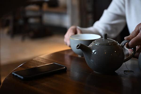 Hands holding a cup of tea and teapot with a mobile phone at a wooden table. Getting ready to phone a friend or phone a counsellor in London | Work Life balance during COVID 19 health crisis | Online therapy in London from YTherapy UK