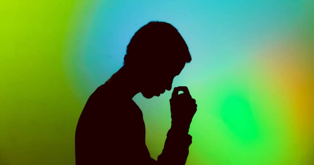A silhouette of a man's profile against a blended blue, green and orange background. The man has lowered his head towards his hand as if feeling stressed and about to burn out. The man is contemplating about stress and burnout therapy.