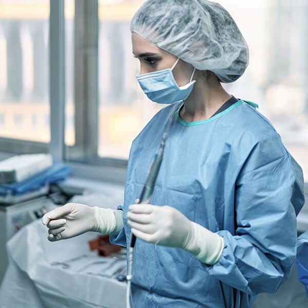 Female NHS surgeon wearing blue gown and gloves prepares for trauma surgery. As a critical care surgeon working in acute care units, this A&E surgeon is often traumatised by the wounded patients she sees.