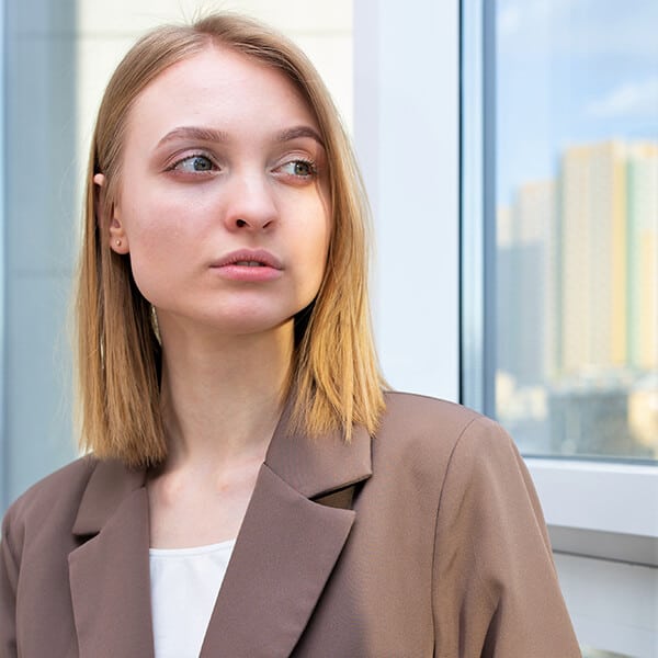 A young professional woman stands in front of a sunlit window. This social worker experiences work-related anxiety and seeks help from YTherapy.