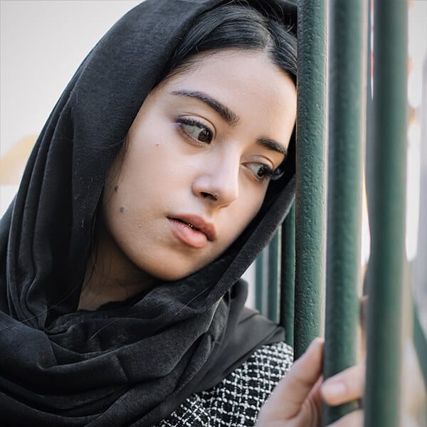 Young woman wearing a hijab headdress leans against fence bars. The woman experiences flashbacks and is looking for therapy for trauma and PTSD in London.