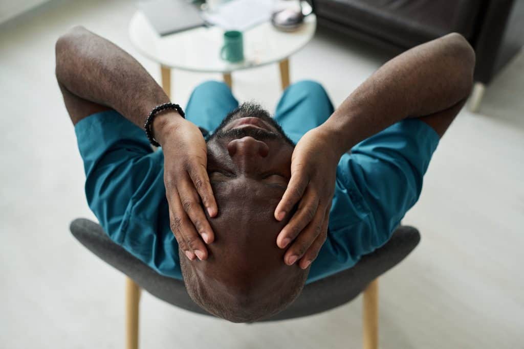 A distressed man sitting back in a chair with his hands pressed against his head, illustrating physical tension and overwhelm often associated with trauma.